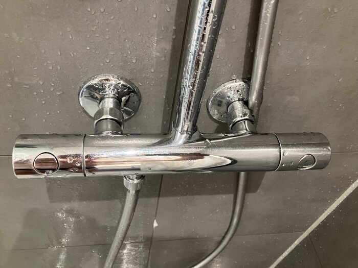 En kromfärgad duschblandare med två vred, monterad på en kaklad vägg, täckt av vattendroppar.