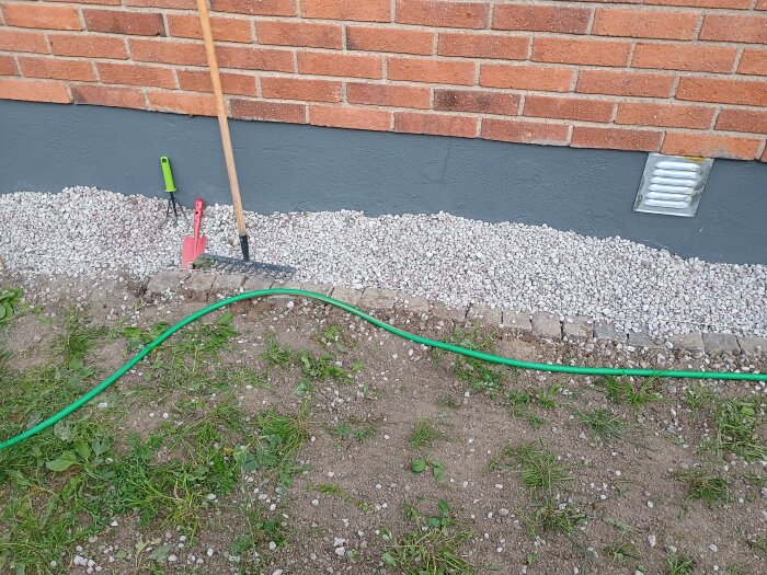 Räfsa, spade och kratta vid marksten och grus längs röd tegelvägg med grön vattenslang liggande ovanför gräs och jord.