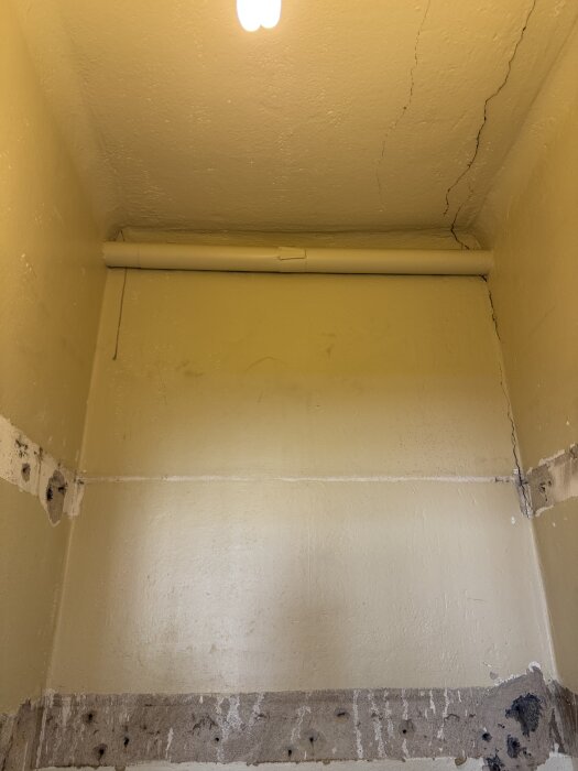 Sprucken vägg och rör bakom rivet material, del av rum som förbereds för badrumsbygge. Oklart föremål, inuti verkar dammfyllt.