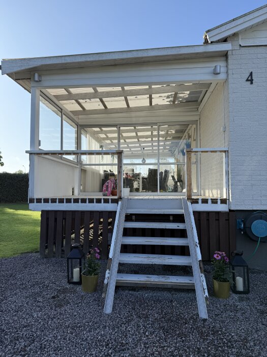 Utbyggd veranda med glaspartier och trappa, omgiven av grus och grönska. Två lyktor och blommor står vid trappan.