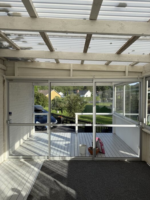 Skjutpartier vid en täckt terrass med utsikt över trädgård och gräsmatta. Inomhus syns ett golv med matta och parkettgolv.