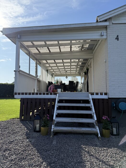 Trappa leder upp till inglasad altan med möbler. Ljus trapp, två lyktor och blomkrukor på grusgång. Soligt väder och klarblå himmel.