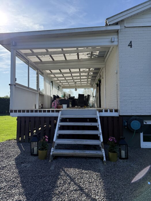 Pergola med vitmålad trappa och skjutpartier, utomhusmöbler syns. Solsken och blå himmel. Trädgård i bakgrunden.