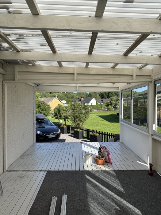 Uteplats med vita träskjutpartier mot en trädgård, grå matta och målningsutrustning. Bil och plank i bakgrunden.