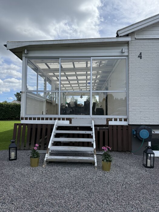 Inglasad veranda med skjutdörrar och en trappa framifrån. Två blomkrukor och två lykta pryder ingången.