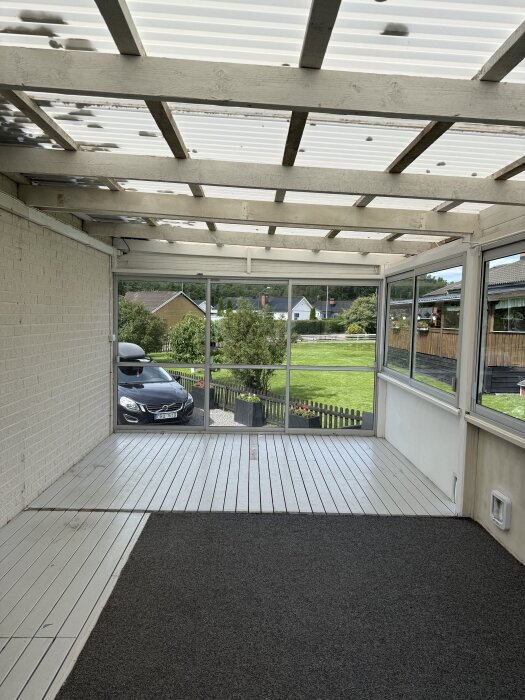 Inglasad veranda med skjutdörrar mot trädgården, delvis mattlagt golv, och utsikt över grönska och parkerad bil framför huset.