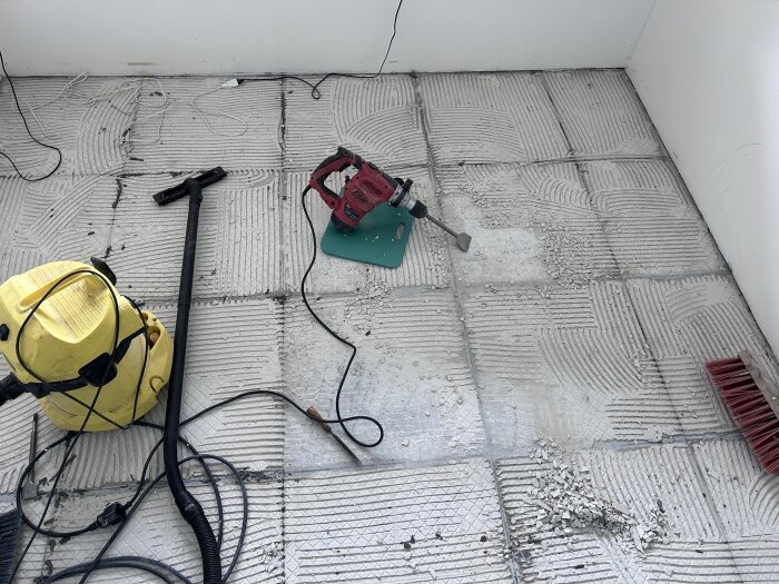 Verktyg för golvrenovering, inklusive en borrmaskin, dammsugare och borste, ligger på ett rum med borttaget kakel, där fixen fortfarande syns på golvet.