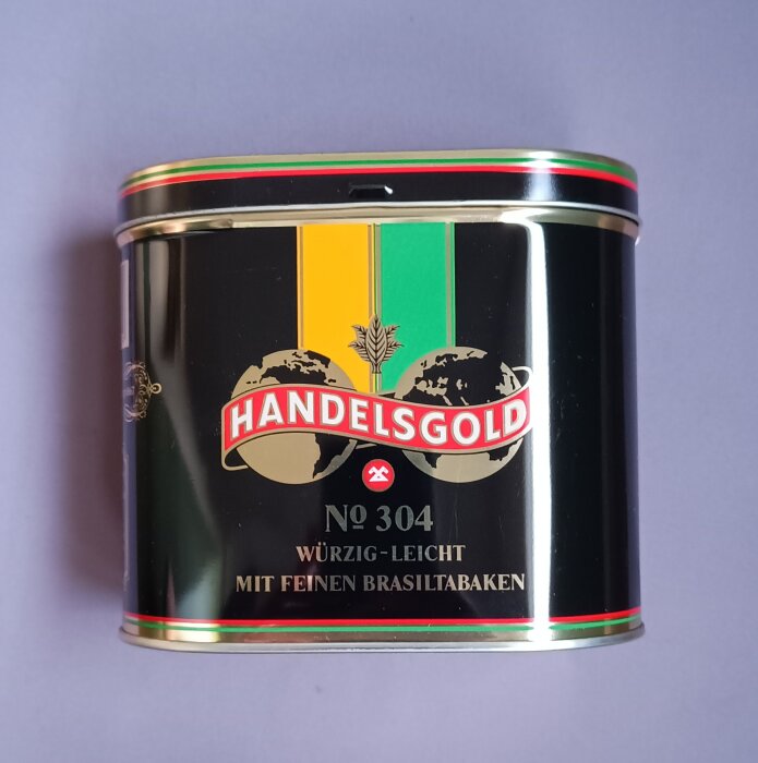 Svart plåtask med texten "Handelsgold No 304, würzig-leicht, mit feinen Brasiltabaken" och en design med jordglob och vete.