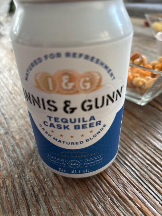 En burk Innis & Gunn Tequila Cask Beer på ett träbord med snacks i bakgrunden.