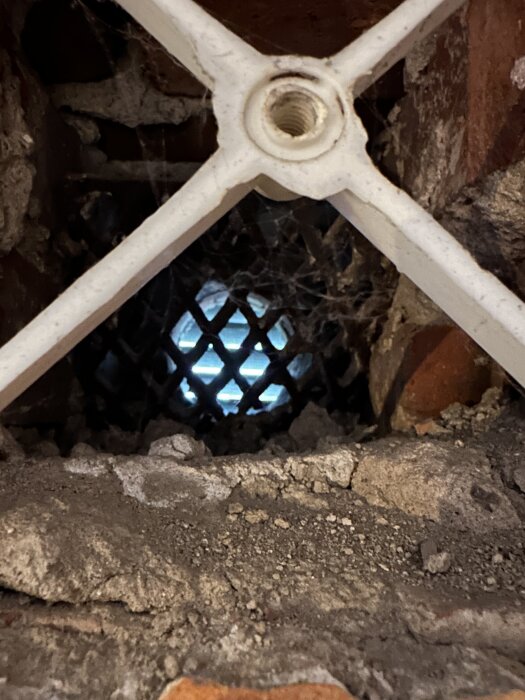 Friskluftsventil i tegelvägg utan rör, synligt hål, omgiven av smuts och spindelnät, med ett justerbart metallgaller.