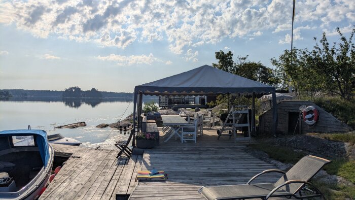 Trädäck vid vattnet med ett tält för skugga, flera stolar och bord under, solstolar och en båt till höger i bilden.