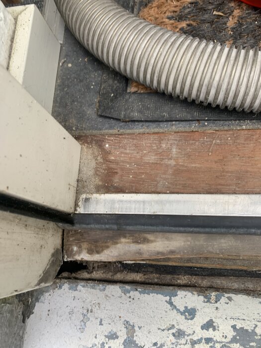 Rutten tröskel vid ytterdörr, omgiven av metallslang och sliten dörrmatta. Tröskeln visar tecken på skada och slitage.