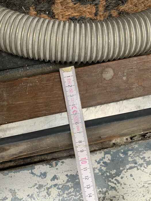 Mätstock mäter höjd på sliten tröskel av trä intill en spiralformad slang och sliten golvyta.