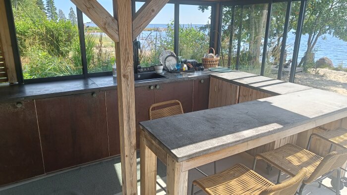 Utekök med träkonstruktion och stenskiva, barstolar och utsikt mot natur och vatten. Diskho och korg med disk på bänken.