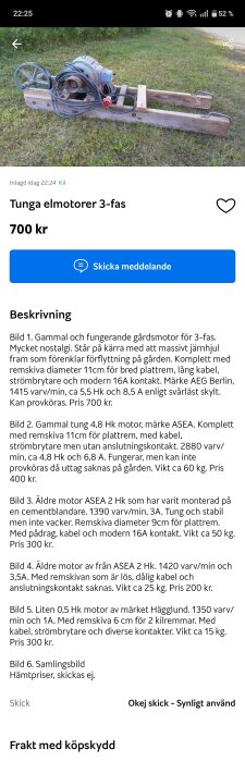 Gammal gårdsmotor på träställning med järnhjul, kablar och remskiva, placerad på gräsmatta. Motor till salu för 700 kr.