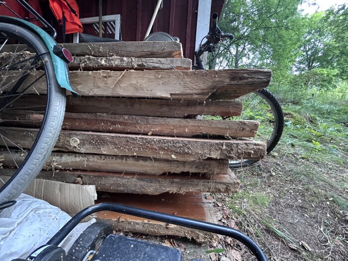 Staplat virke utomhus med grova träplankor, delvis täckt av cykeldelar. Bakgrunden visar grönska och en del av en byggnad.