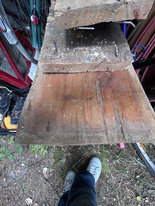Gamla träplankor på varandra, delvis skadade med bortomring av spikar, placerade ovanpå jordig mark intill cykelhjul och verktyg.