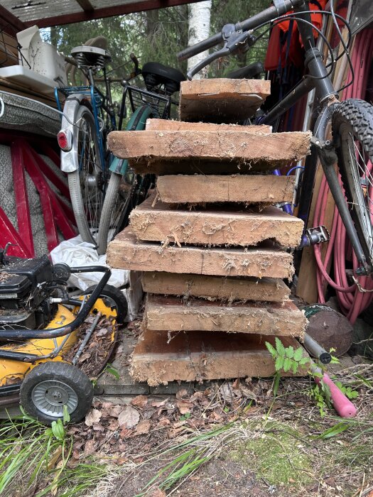 En hög av gamla träbrädor staplade på varandra med cyklar och en gräsklippare i bakgrunden i ett förråd.