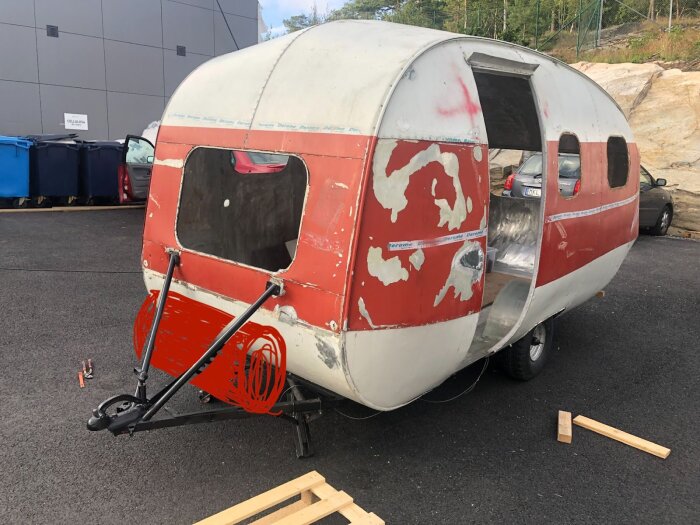 Röd och vit äggformad husvagn under konstruktion, med öppet hål och verktyg runt omkring, står utanför vid en byggnad.