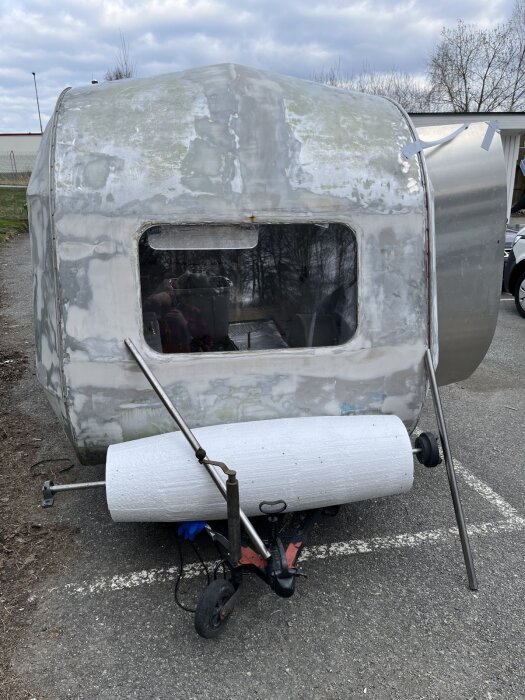 En husvagn under renovering med en cylindrisk konstruktion av cellplast vid fronten, placerad på en parkeringsplats.