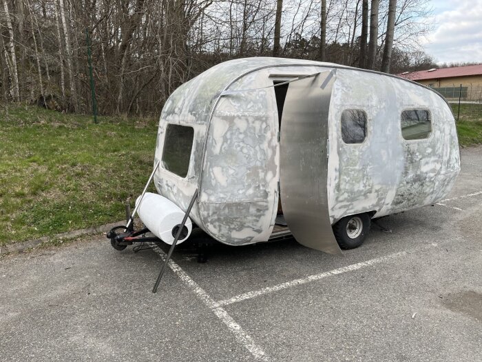 Äggformad husvagn med öppen dörr, sett från sidan. Cylinderformad föremål med plastfolie är fäst framtill. Husvagnen är omgiven av träd och gräsmatta.