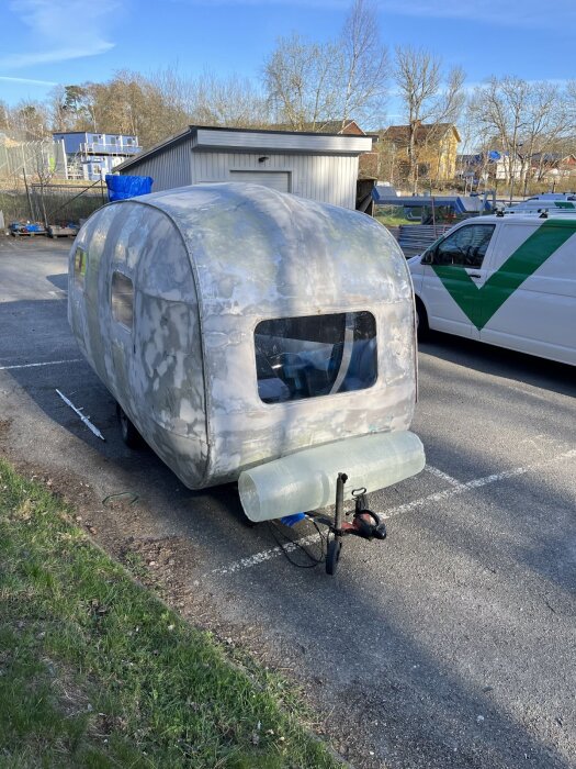 Husvagn med rundad, hemmagjord cylinder framtill för batteriförvaring, stående på en parkeringsplats bredvid en vit skåpbil.