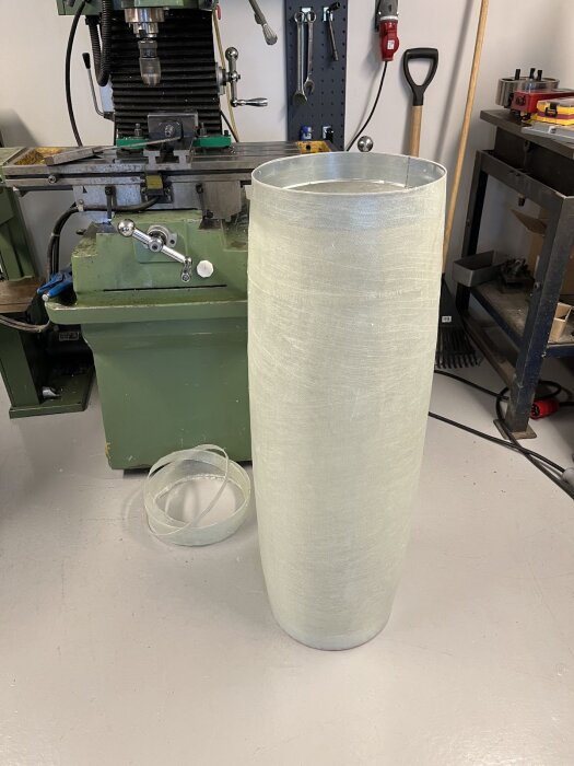 Glasfiberförstärkt cylinderformad batteribox under konstruktion, placerad framför en grön maskin i verkstad.