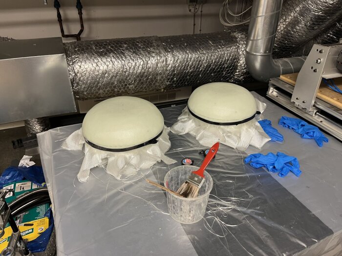 Två halvcirkulära plastdetaljer på ett bord, omgivna av skyddshandskar, pensel och mätband. Bakgrunden visar kanalrör i metall.