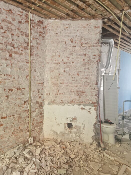Delvis riven tegelvägg med rester av murbruk och spill på golvet, synlig toalett och verktyg, planerad renovering av skorsten.