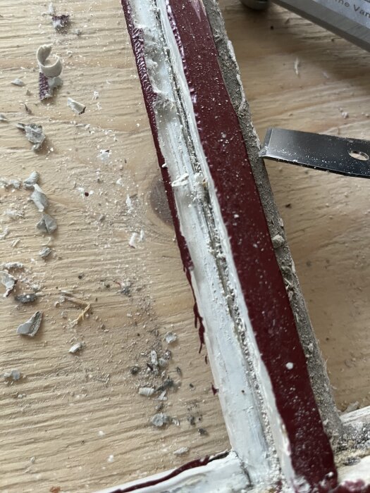 Fönsterkarm med borttaget kitt och rödfärg, verktyg används för att skrapa bort rester; träflisor och kitt syns på ytan.