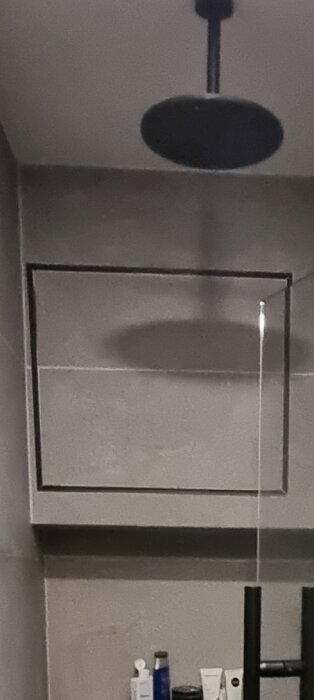 En duschvägg med en fyrkantig lucka med svart ram, inuti finns ventilation. Luckan är fäst med magneter ovanpå kakel.