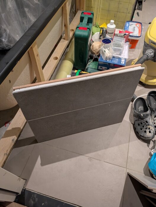 Badrumslucka av kakel lutad mot vägg, omgiven av verktyg och byggmaterial, del av pågående renoveringsprojekt för att dölja ventil bakom.