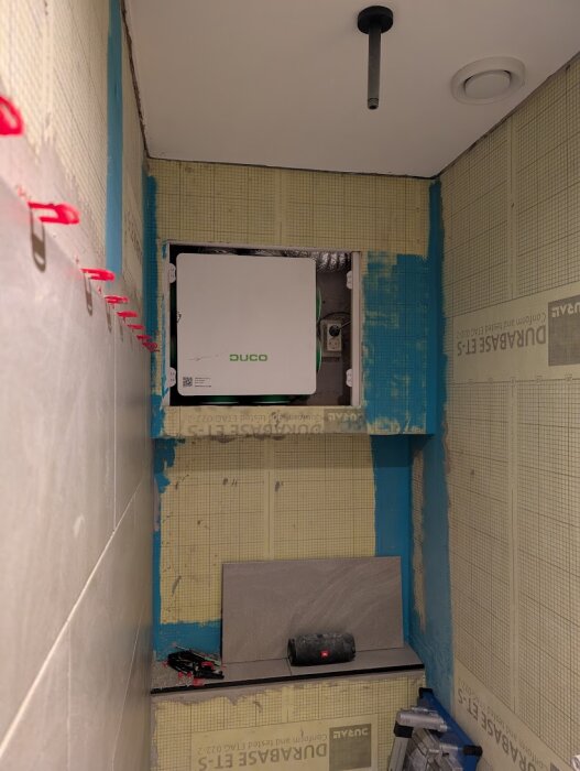 Hål i badrumsvägg avslöjar ventilationssystem med lucka märkt "DUCO", omgivet av blottad konstruktion och isolering.