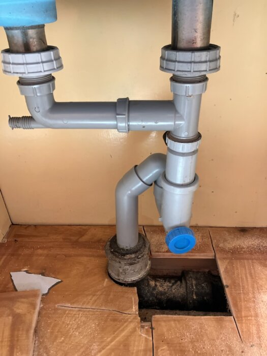 Rörsystem under köksvask med plastkopplingar och synligt gjutjärnsrör, omgivet av sågad träplatta, möjlig plats för läcka indikeras.