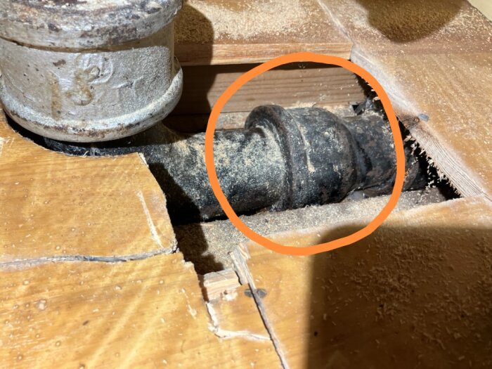 Gjutjärnsrör under köksvasken med vattenläcka markerad med orange cirkel, omgivet av sågspån på träplatta.