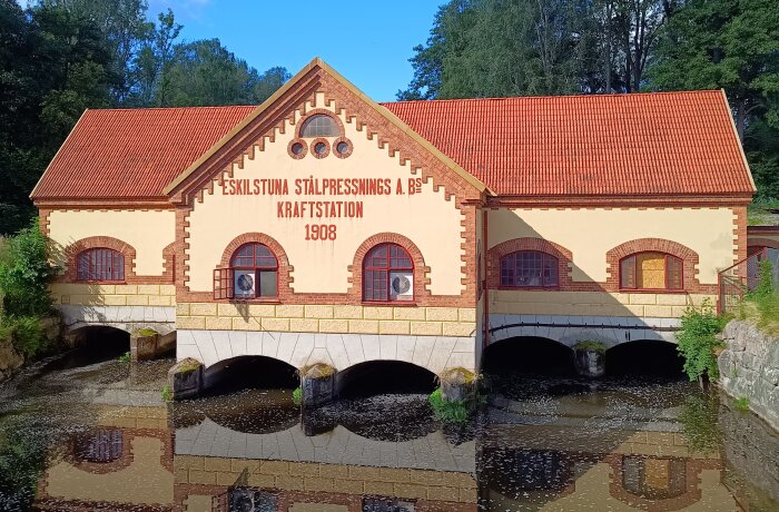 Kraftstationen i Skogstorp, Eskilstuna, från 1908, med originalkomponenter och tydlig arkitektur, reflekteras i vattnet framför.