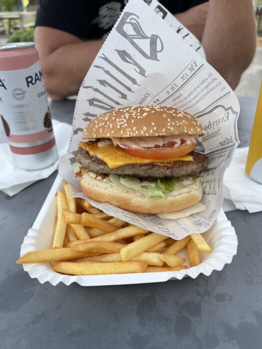 Burger på en bricka med pommes frites, inlindad i papper. Sesambröd, sallad, tomat och ost. Ramona dryckeburk i bakgrunden.