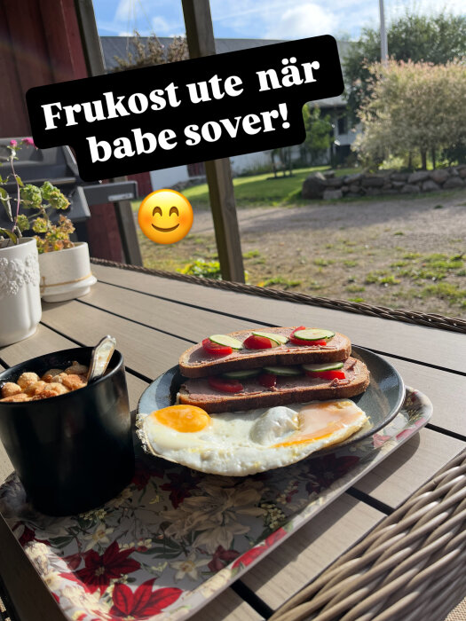 Frukostbricka utomhus med smörgåsar, stekt ägg och flingor bredvid en blomkruka och utemöbler i soligt väder.