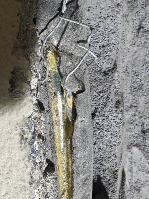Rör med gul isolering i ett vägguttag, omgivet av sprucken betong och metalltrådar.