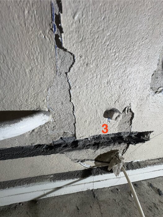 Skadat väggparti med exponerade rör och övermålad mutter markerad med siffran 3, hål i vägg visar byggnadens struktur.