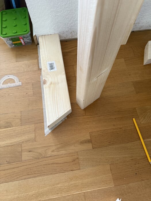 Träreglar på golv, en med avfasad kortsida, bredvid en lodrät regel. Blyertspenna och plastvinkel på parkettgolv.