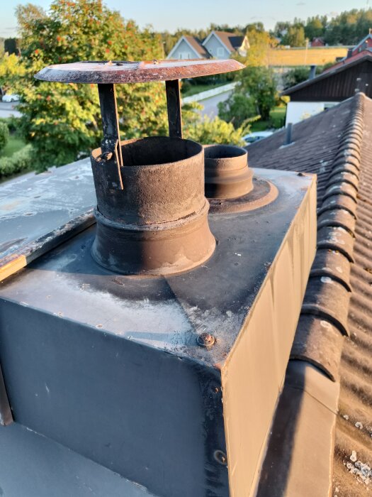 Skorsten med två ventilationsrör, ett större med hatt och ett mindre utan lock, på taket av ett hus i ett grönskande område.