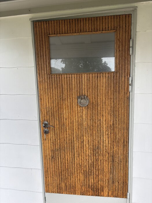 Brun trädörr med glasruta upptill och omgivande vit eternitfasad.