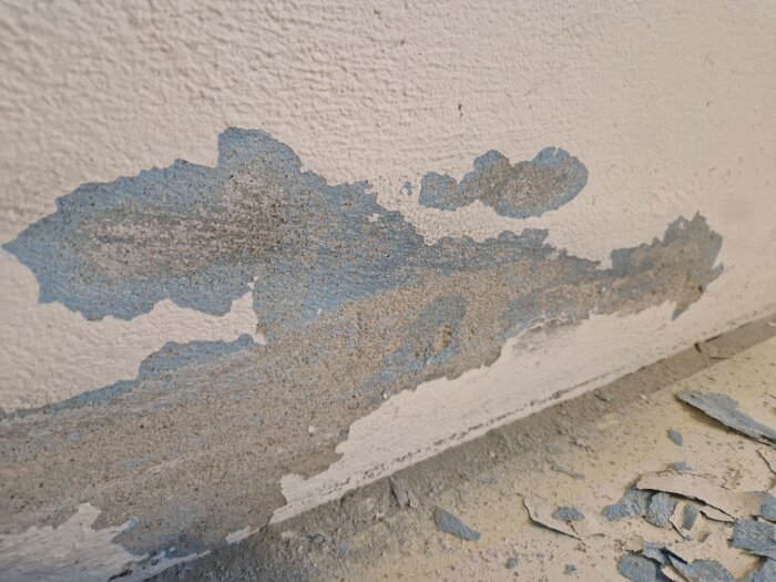 Blå färgfläckar och avflagnad puts på en väggyta, med färgrester på golvet.