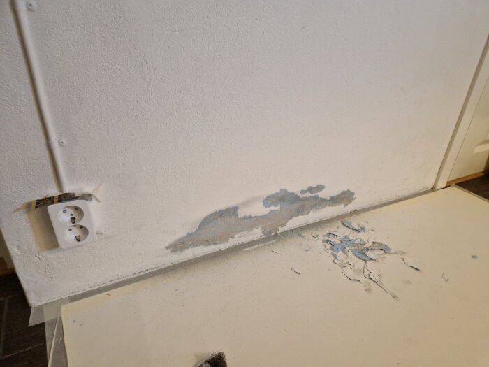 Del av vit vägg med avskavd färg och en fläck vid ett dubbeluttag. Golvet täckt med papper och färgflagor synliga.