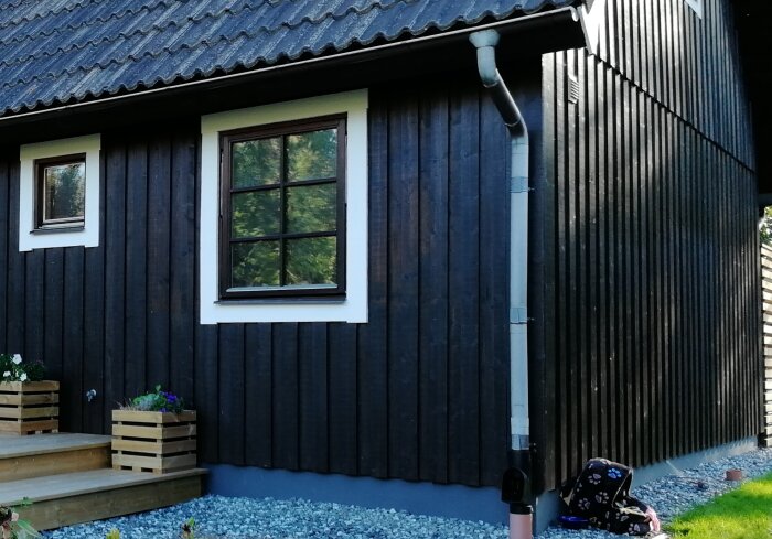 Svartmålad husvägg med vita fönsterkarmar och betongpannetak. Grusbädd vid husets fot och planteringslådor vid trappan.