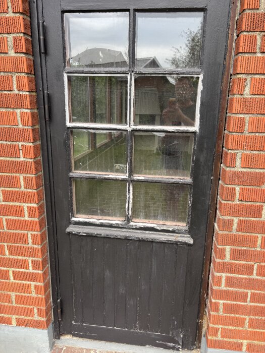 Svart trädörr med glasrutor och röd tegelvägg i vinterträdgård under renovering, spegling av person i glaset.