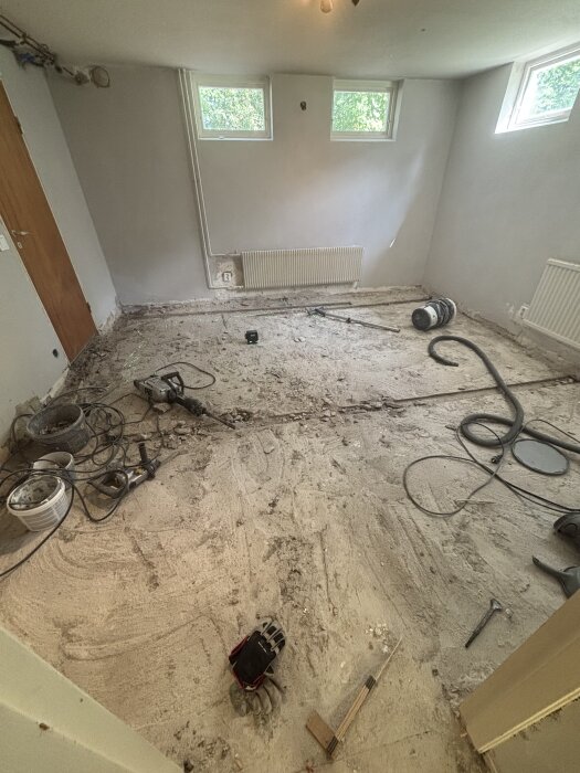Rivningsprojekt i källarrum med blottat betonggolv, verktyg och byggmaterial utspridda, inklusive borrmaskin och dammsugarslang.