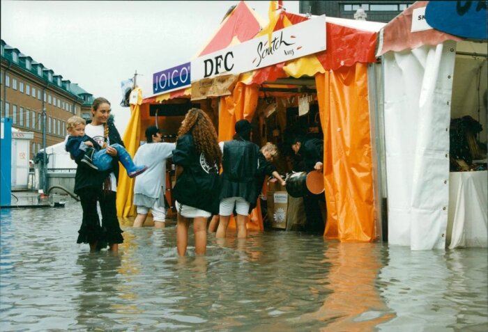 Personer vadar genom översvämning vid tält med skyltar för Joico och DFC, en kvinna bär ett barn.