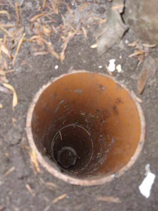 Brun spolbrunn med borttappat lock sedd ovanifrån, omgiven av jord och löv.